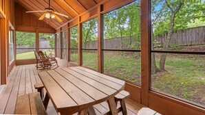 Eine überdachte Terrasse mit Holztisch und -stühlen, großen Fenstern und einem Deckenventilator.