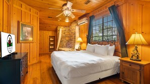 Ein Schlafzimmer in einer Holzhütte mit einem großen Bett, einem Deckenventilator, einer Steinwand und einem Fenster mit Vorhängen.
