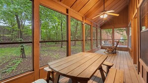 Una terraza de madera con una mesa y bancos, amplias ventanas que dan a una zona boscosa.