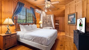 Un dormitorio de cabaña de madera con cama, mesitas de noche, ventilador de techo, pared de piedra y un cuadro enmarcado.
