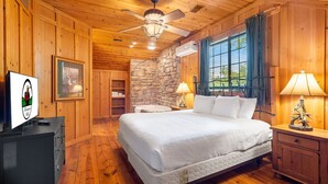 A wooden cabin-style room with a large bed, a ceiling fan, a stone wall, and a window with curtains.