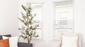 A living room with a grey sofa, a white armchair, two woven ottomans, a tall plant, and white-framed windows with blinds. - CrestNest- Modern 2BD, 1BA - 2.5 Blocks to Beach (Wildwood)