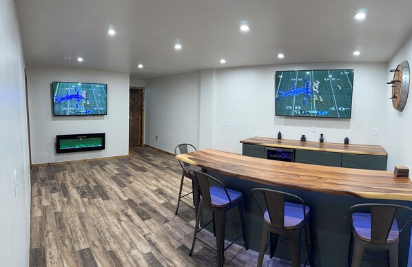 A modern kitchen with a wooden bar, two TVs mounted on the wall, and a wooden floor. - 4-bed Cabin- ATV/UTV trail access from the driveway with a bar/covered patio (Crivitz)
