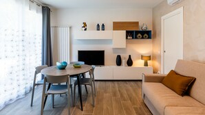 Ein modernes Wohnzimmer mit einem runden Esstisch, zwei Stühlen, einem beigen Sofa und einem Fernseher auf einem weißen Schrank.