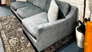 A living room with a grey sofa, a round wall clock, a potted plant, and a patterned rug. - `The Hideaway` Modern Apartment w/Sauna and Grill (Troy)