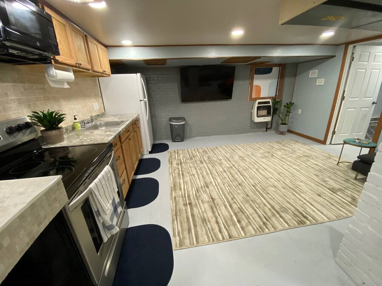 Una cocina moderna con armarios de madera, un microondas negro, una estufa y un lavamanos. Hay una alfombra grande en el suelo y un cubo de basura pequeño en la esquina.
