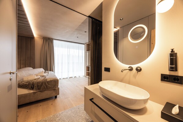 Una habitación de hotel moderna con una cama grande, un espejo redondo con luz, un lavabo blanco y una botella de champú.