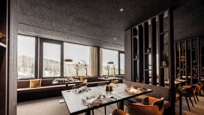 A modern dining area with a long table, multiple chairs, and a seating area with a view of a snowy landscape. - Alma Alpina Lodge (Nova Ponente)
