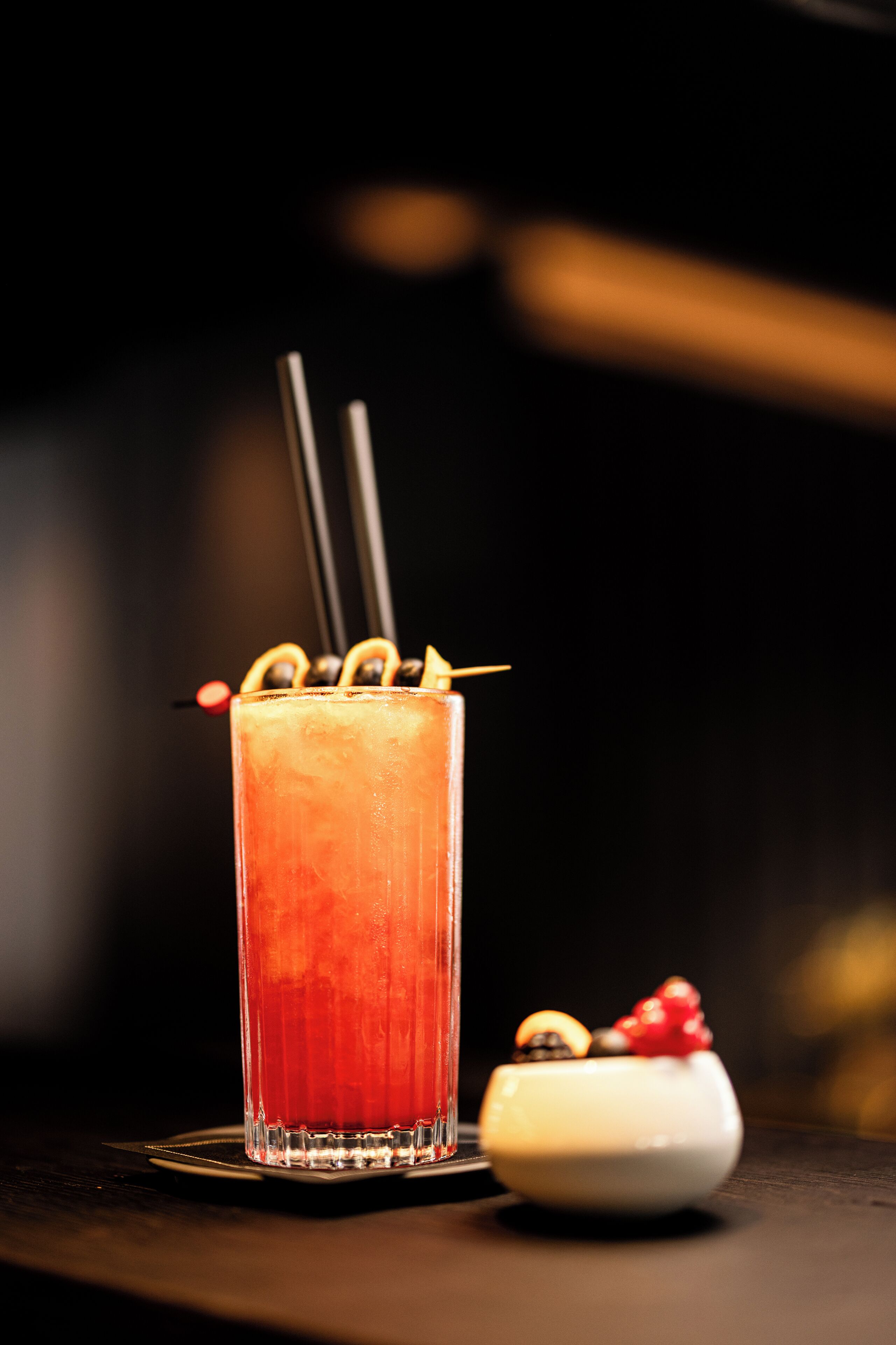 A tall glass with a red beverage, garnished with lemon slices and a straw, accompanied by a small white dish with a red fruit.