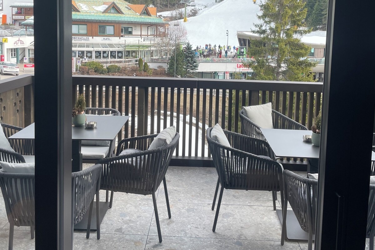 Un balcone con sedie in vimini e un tavolo, con vista su una pista da sci e edifici.