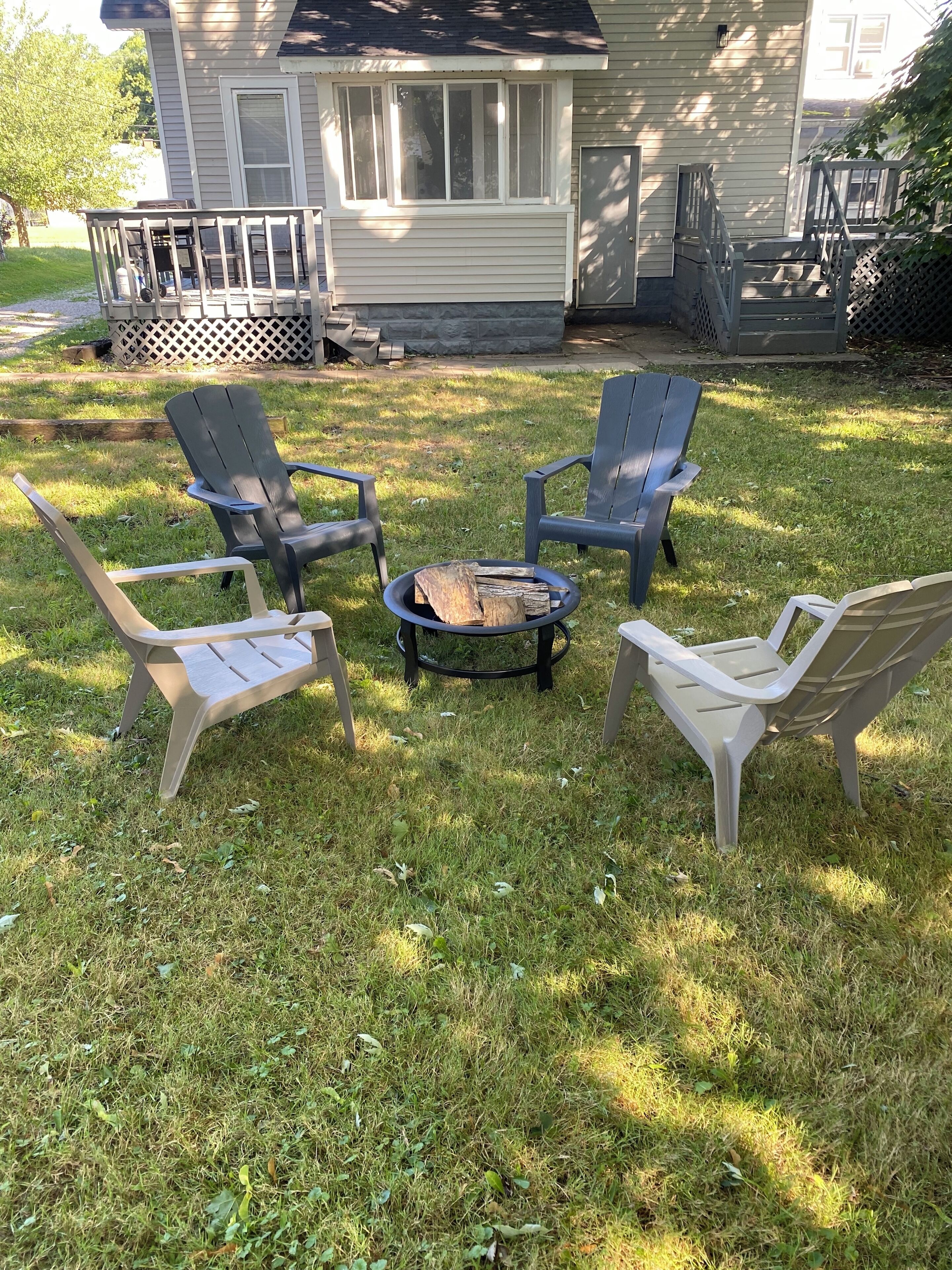 Un jardin avec un foyer entouré de quatre fauteuils Adirondack, une terrasse en bois et une maison en arrière-plan.