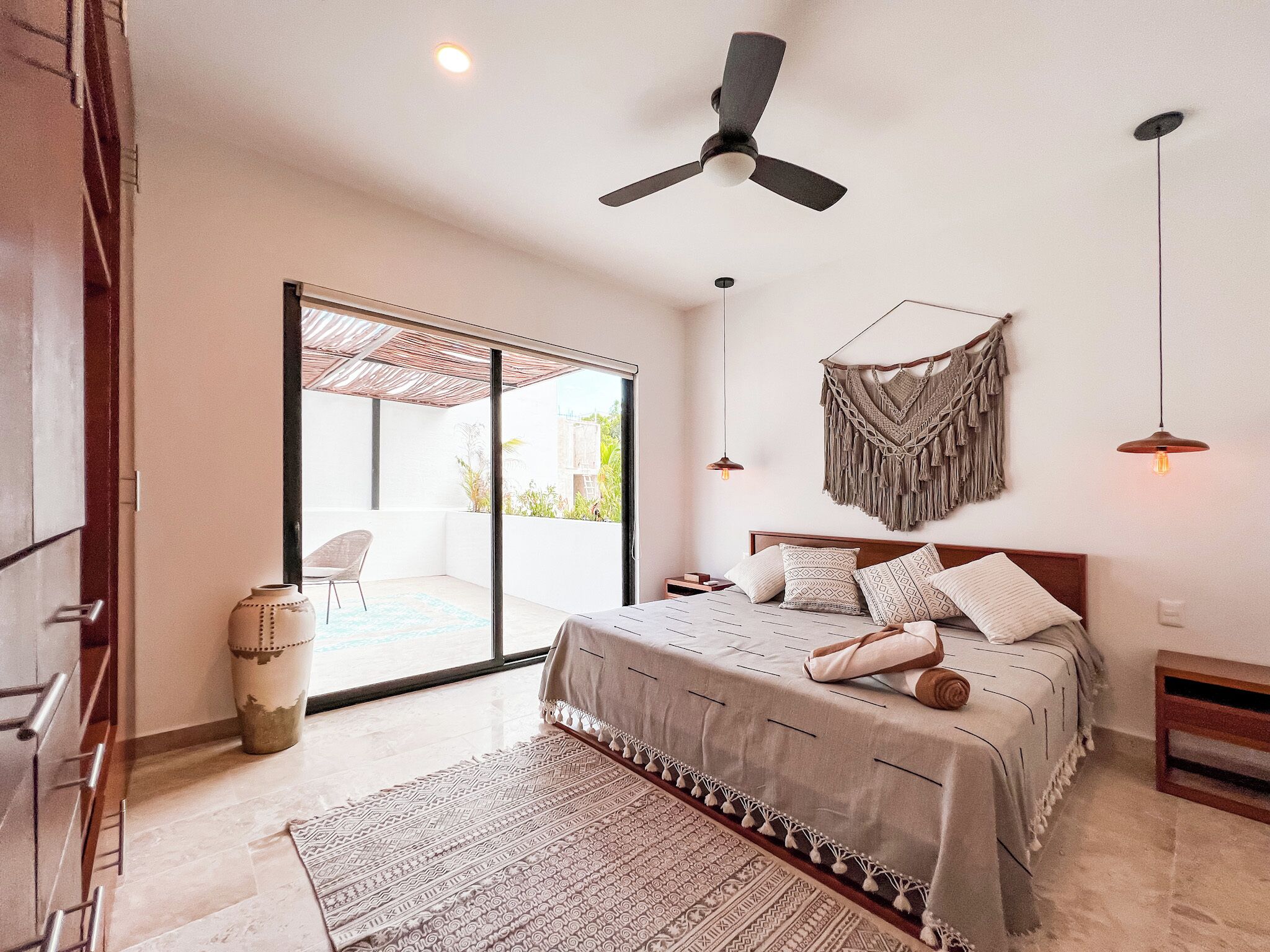A bedroom with a bed, a ceiling fan, a macrame wall hanging, and a view through a sliding glass door to an outdoor area.