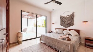 A bedroom with a bed, a ceiling fan, a macrame wall hanging, and a view through a sliding glass door to an outdoor area.