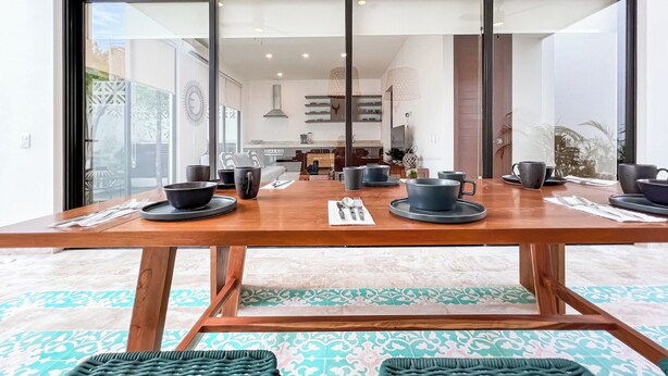 A dining area with a wooden table set for four, a large window with a view of the kitchen, and a patterned floor.