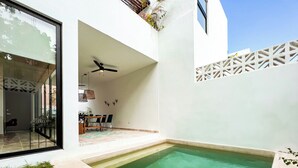A modern outdoor pool area with a ceiling fan, a dining table with chairs, and a small garden.