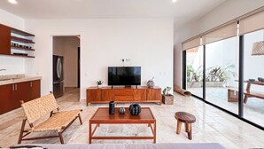 A modern living room with a flat-screen TV, wooden furniture, a dining area with a bench, and a kitchen with a refrigerator.