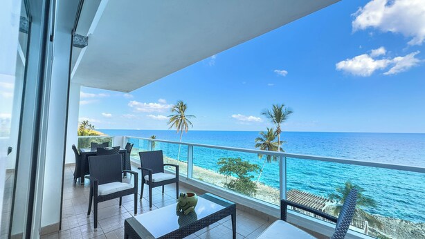 A balcony with a view of the ocean, furnished with chairs and a table, and a glass railing.