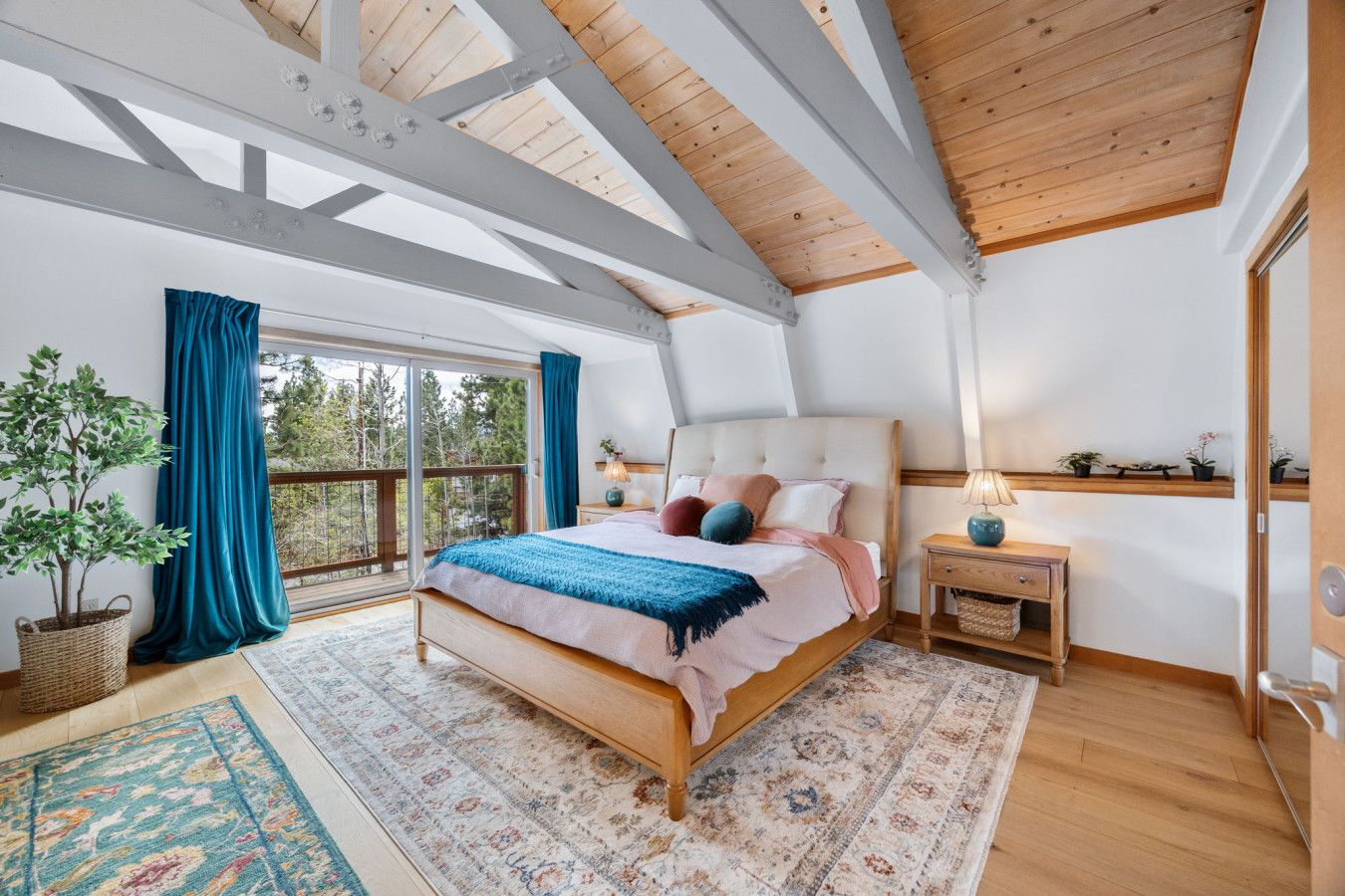 A bedroom with a large bed, a nightstand, a lamp, a potted plant, and a view through a window.