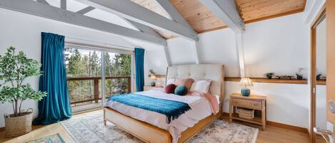 A bedroom with a large bed, a nightstand, a lamp, a potted plant, and a view through a window.