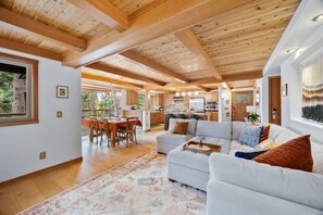 Amplia sala de estar con un sofá grande de color gris, un comedor con sillas de madera, y una cocina con vigas de madera y electrodomésticos modernos.