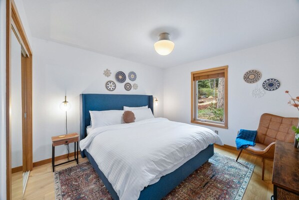 Un dormitorio con una cama grande, una mesita de noche de madera, una silla, una ventana con vistas a zonas verdes y decoración mural.