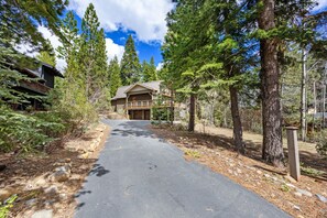 A cabin with a paved driveway leading to a wooden deck and garage, surrounded by tall pine trees.