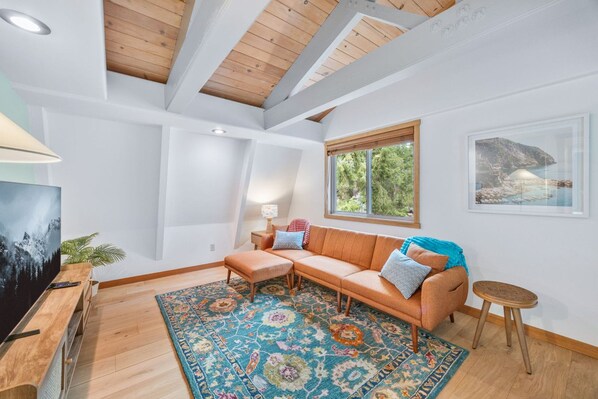 A modern living room with a wooden ceiling, a large window with a view of trees, a sofa with cushions, a small table, and a patterned rug.