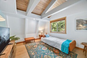 Een slaapkamer met een bed, een bankje, een bureau met een televisie, een raam met uitzicht op bomen en een kamerbreed tapijt met dessin.