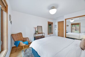Un dormitorio con una cama grande, una silla de madera, una mesita con un florero y un espejo en la pared.