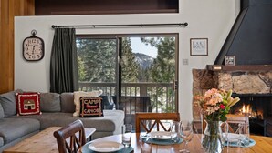 Una sala de estar acogedora con chimenea, una mesa de comedor para cuatro personas y un amplio ventanal con vistas a un bosque.