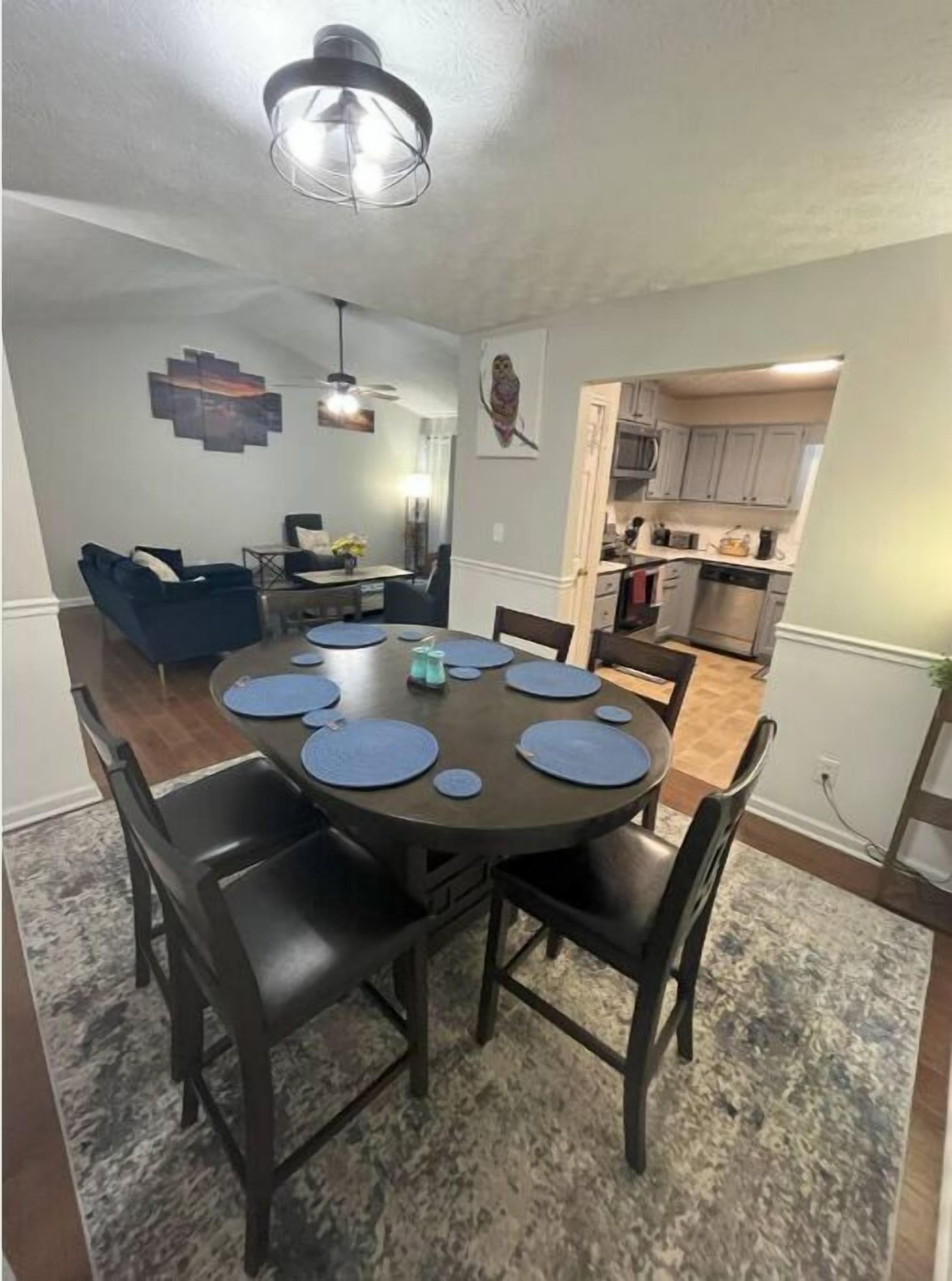Un coin repas avec une table ronde, des chaises foncées et des sets de table bleus. Un luminaire est suspendu au plafond au-dessus de la table.
