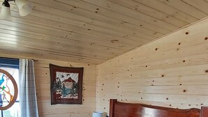 Habitació de cabana de fusta amb un llit, una tauleta de nit, una finestra amb cortines i un penjoll a la paret.