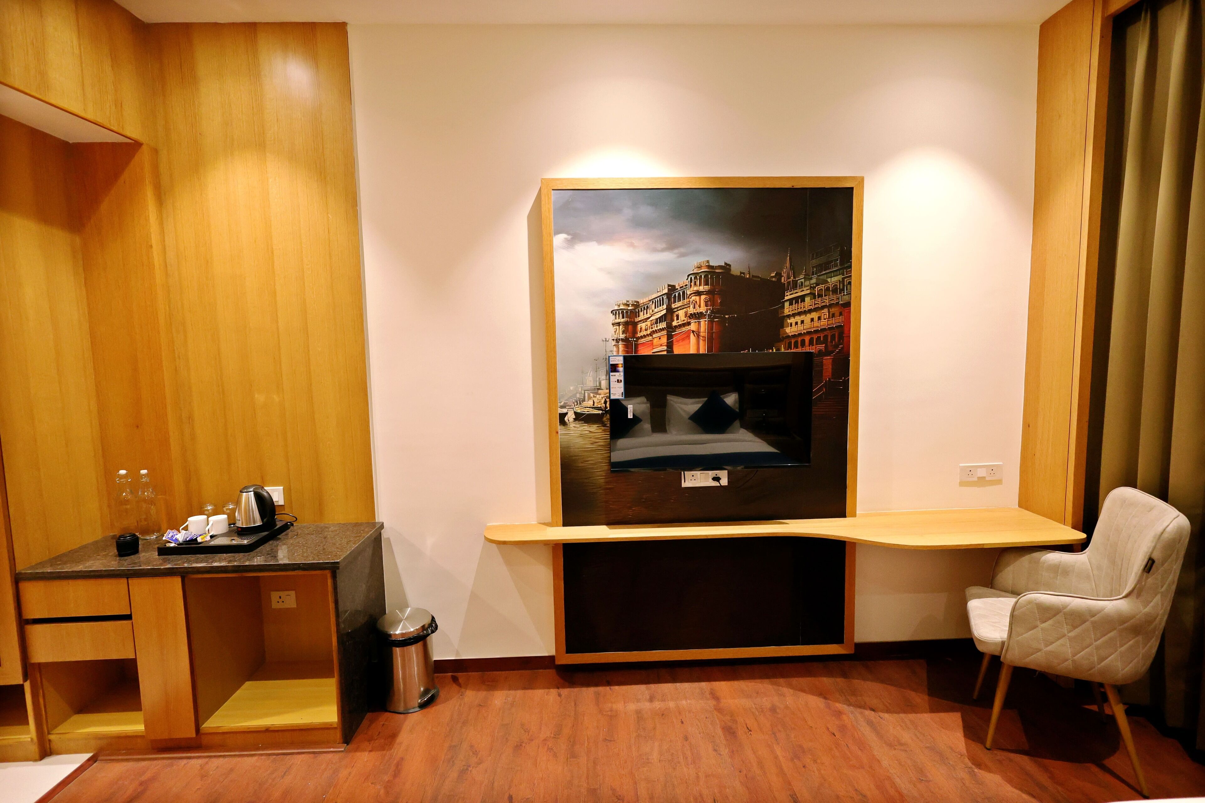 A compact hotel room with a wooden desk, a chair, a TV mounted on the wall, and a trash bin.
