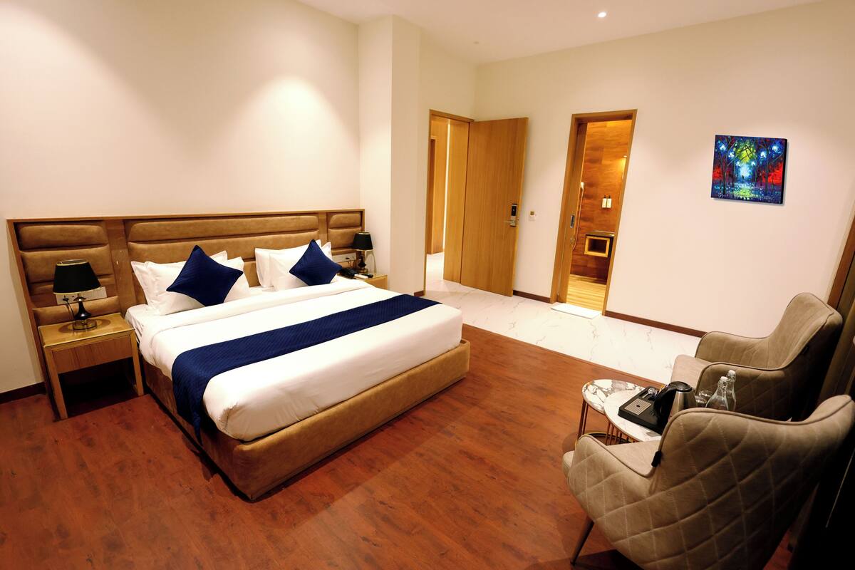 Una habitación de hotel con una cama grande, una mesita de noche con lámpara, una silla y un cuadro en la pared.