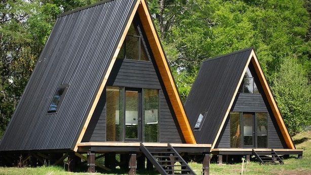Deux cabanes modernes en bois, de forme triangulaire et dotées de grandes fenêtres, sont installées dans une zone herbeuse, avec des arbres en arrière-plan.