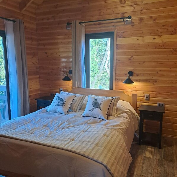 A wooden cabin bedroom with a bed, two bedside lamps, and a window with curtains. - Alto Terralis (Pucón)
