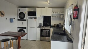 A compact kitchen with a washing machine, microwave, oven, and a small dining area with two stools. - Orewa Kiwi Cabins - Kereru Cabin (Orewa)