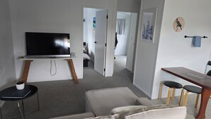 A modern living room with a flat-screen TV on a stand, a sofa, a small dining table with stools, and a hallway leading to another room. - Orewa Kiwi Cabins - Kereru Cabin (Orewa)