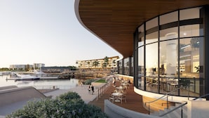 A waterfront restaurant with outdoor seating overlooking a marina, featuring a modern architectural design with large glass windows and wooden deck.