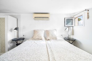 Un dormitorio con cama, dos mesitas de noche, aire acondicionado instalado en la pared, ventana con vistas y una pequeña mesa con lámpara.