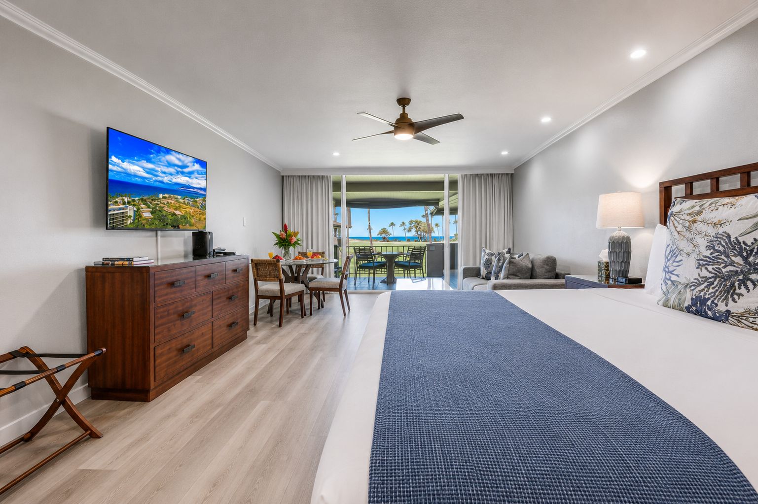 A hotel room with a bed, a desk, a television, a ceiling fan, and a view of the ocean.