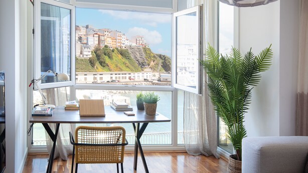 A room with a view of a coastal town, a desk with books and a plant, and a wicker chair.