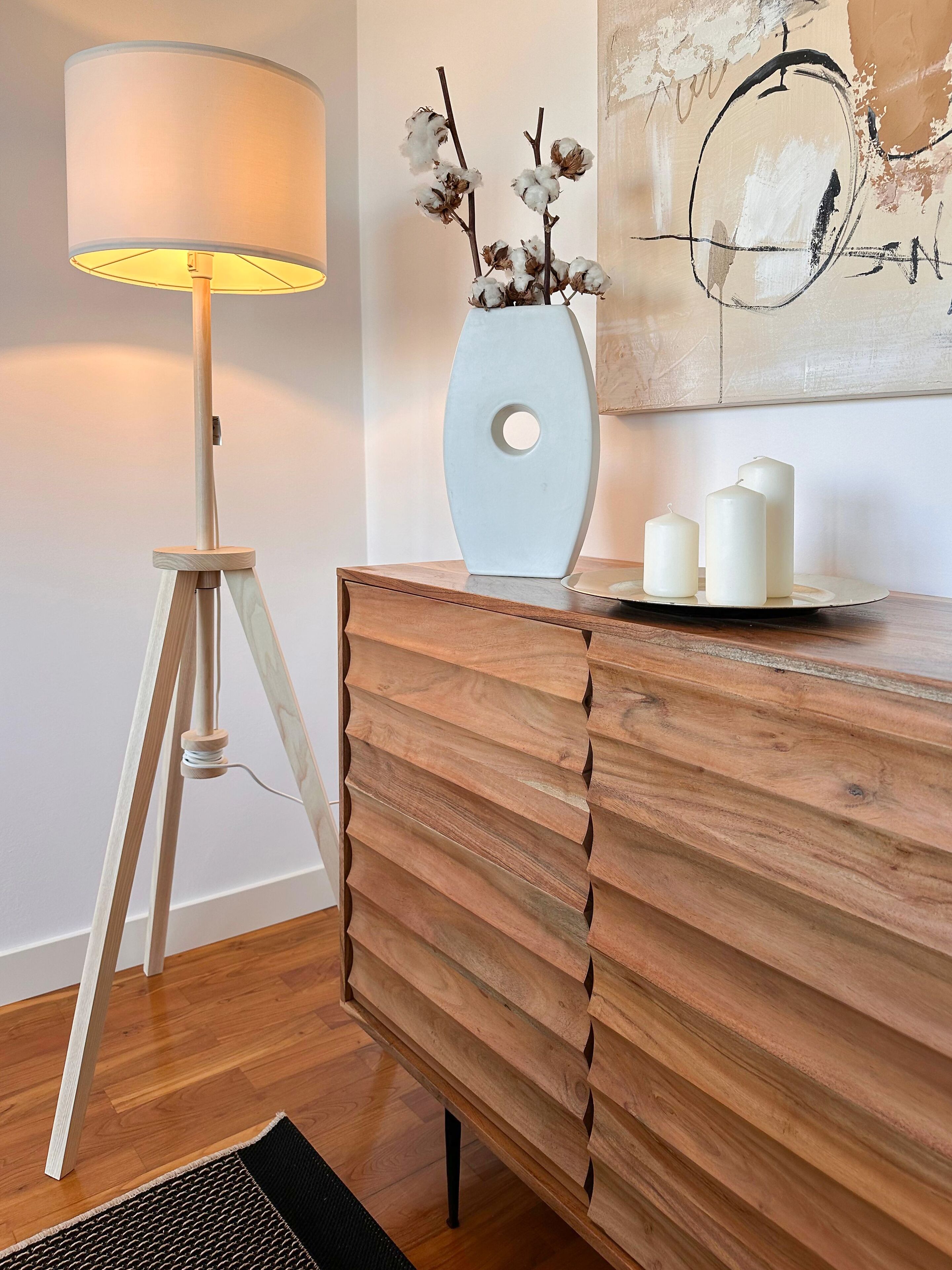 Een houten dressoir met een vaas gedroogde bloemen en kaarsen, een lamp en een schilderij aan de muur.