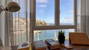 A room with a large window overlooking a coastal view, a desk with books, a lamp, and a potted plant.