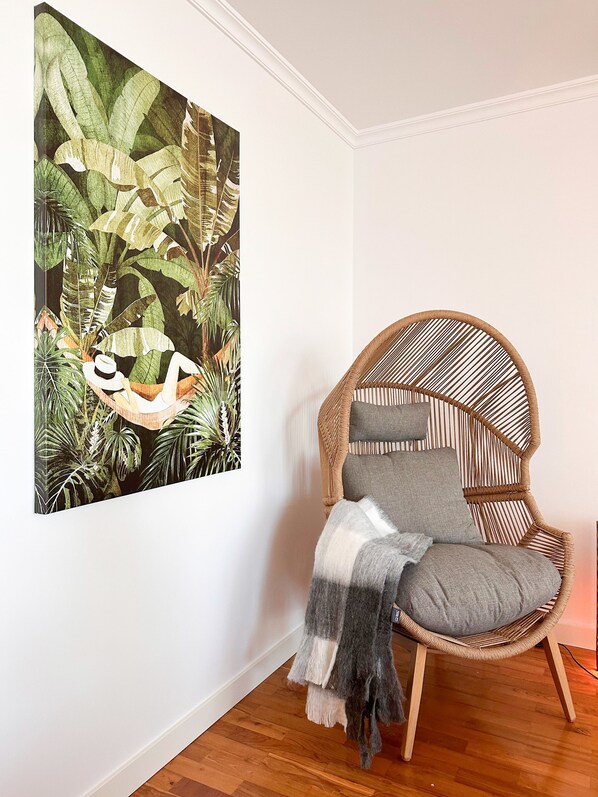 A room with a large botanical print on the wall, a wicker chair with cushions, and a wooden floor.