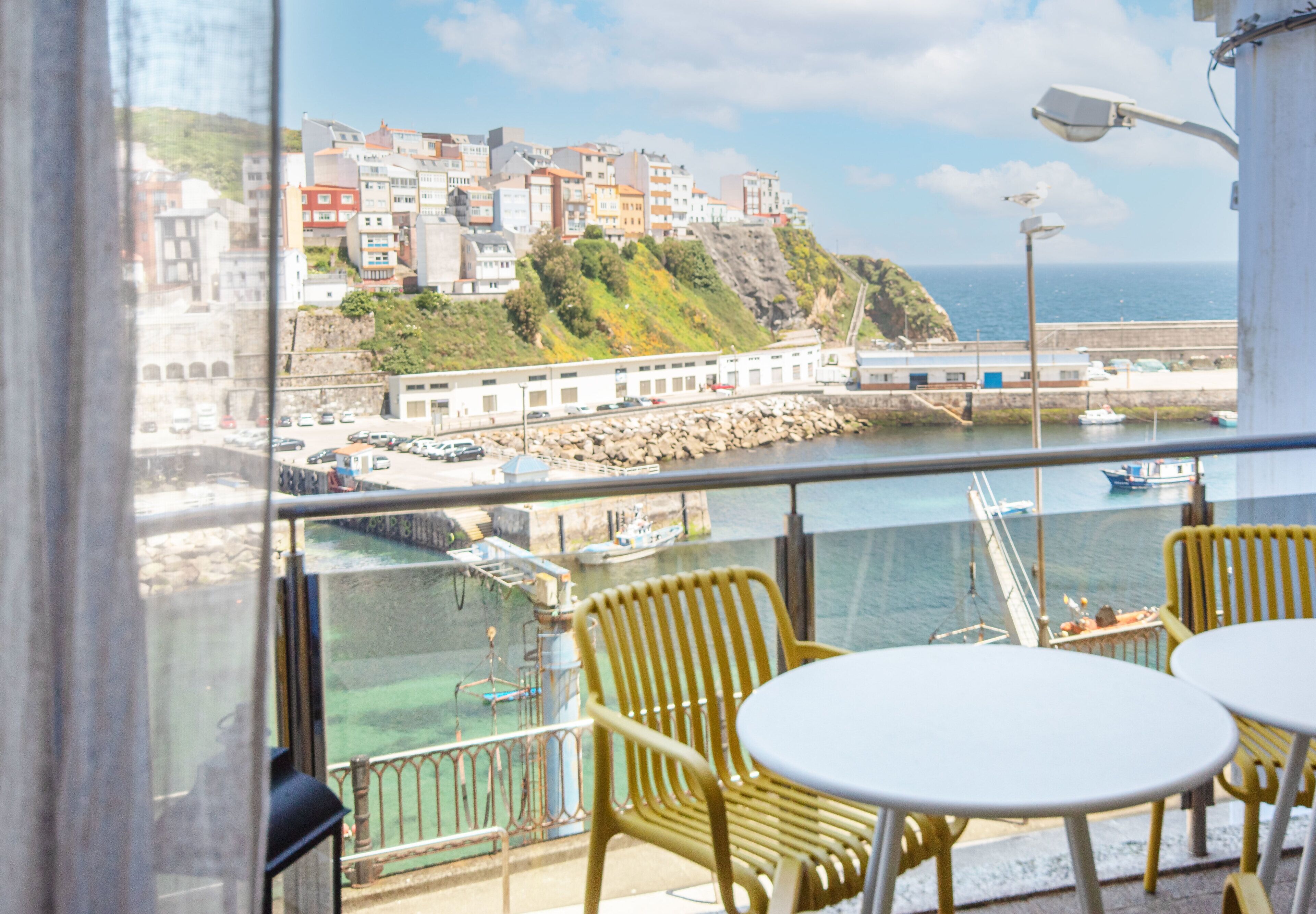 A balcony with yellow chairs and a table, overlooking a coastal town with buildings on a hillside and boats in the water.