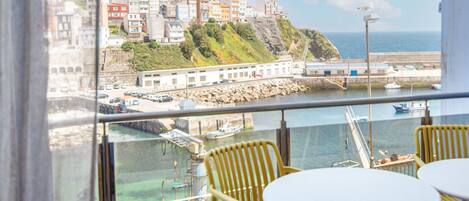 A balcony with yellow chairs and a table, overlooking a coastal town with buildings on a hillside and boats in the water.