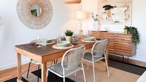 A dining area with a wooden table, white wicker chairs, a woven pendant light, a round mirror, and a minimalist painting on the wall.
