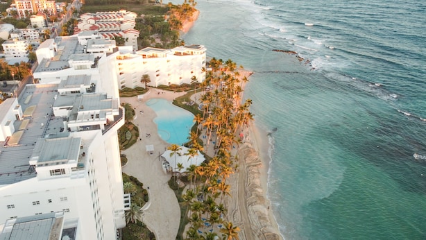 An aerial view of a coastal hotel with a swimming pool, beachfront, and palm trees.