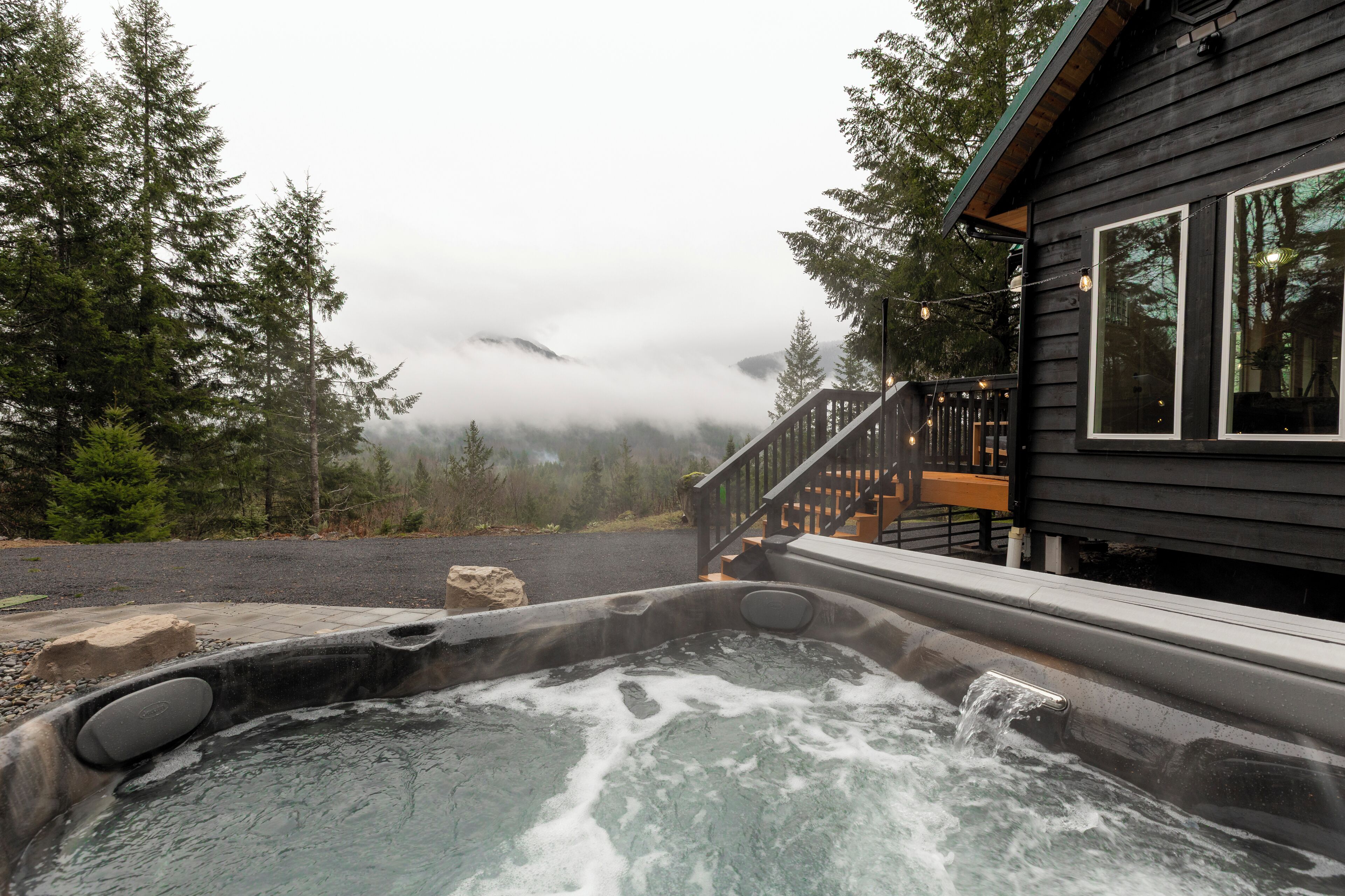 A hot tub with a wooden deck and a staircase leading to a cabin surrounded by trees and fog.
